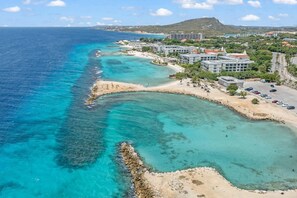 Un resort costero con villas frente a la playa, aguas turquesas cristalinas y un paisaje montañoso de fondo.