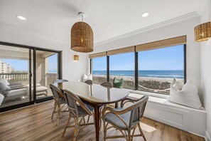 Ein Essbereich mit Tisch und Stühlen, große Fenster mit Blick auf den Strand und eine Pendelleuchte aus Korbmaterial.