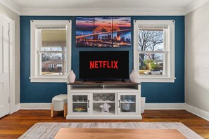 Ein Wohnzimmer mit an der Wand montiertem Fernseher, einem Fenster mit Blick auf eine Brücke und einer weißen Unterhaltungsecke darunter.