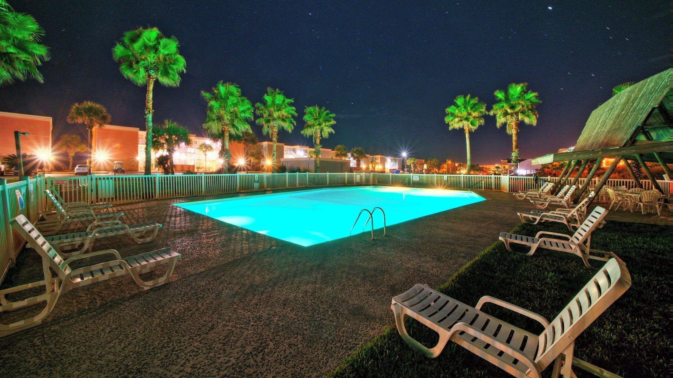A swimming pool with lounge chairs, palm trees, and a night sky.