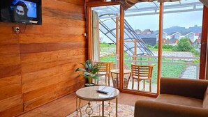 A cozy room with wooden walls, a television, a round table with chairs, a sofa, and a large window with a view of greenery and buildings. - Cabin Dieng Db (Dieng Kulon)