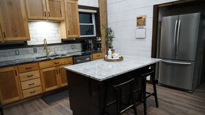 A kitchen with wooden cabinets, a stainless steel refrigerator, a marble countertop, and a bar stool. - The Barn at Reed | Hot Tub + Horse Friendly (Reeds)