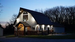 A large barn-style building with a prominent gable roof, a stone foundation, and a porch with a wooden railing. - The Barn at Reed | Hot Tub + Horse Friendly (Reeds)
