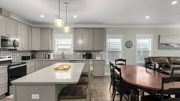 A modern kitchen with grey cabinets, a central island, a dining area with a wooden table and chairs, and pendant lighting.