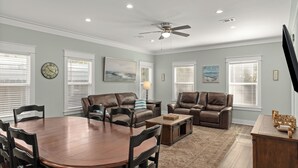 A spacious living room with a dining table, two leather sofas, a coffee table, and a ceiling fan. - Central Pines by Avantstay 30A Wooded Retreat Hot Tub + Private Yard Near Beaches (Santa Rosa Beach)