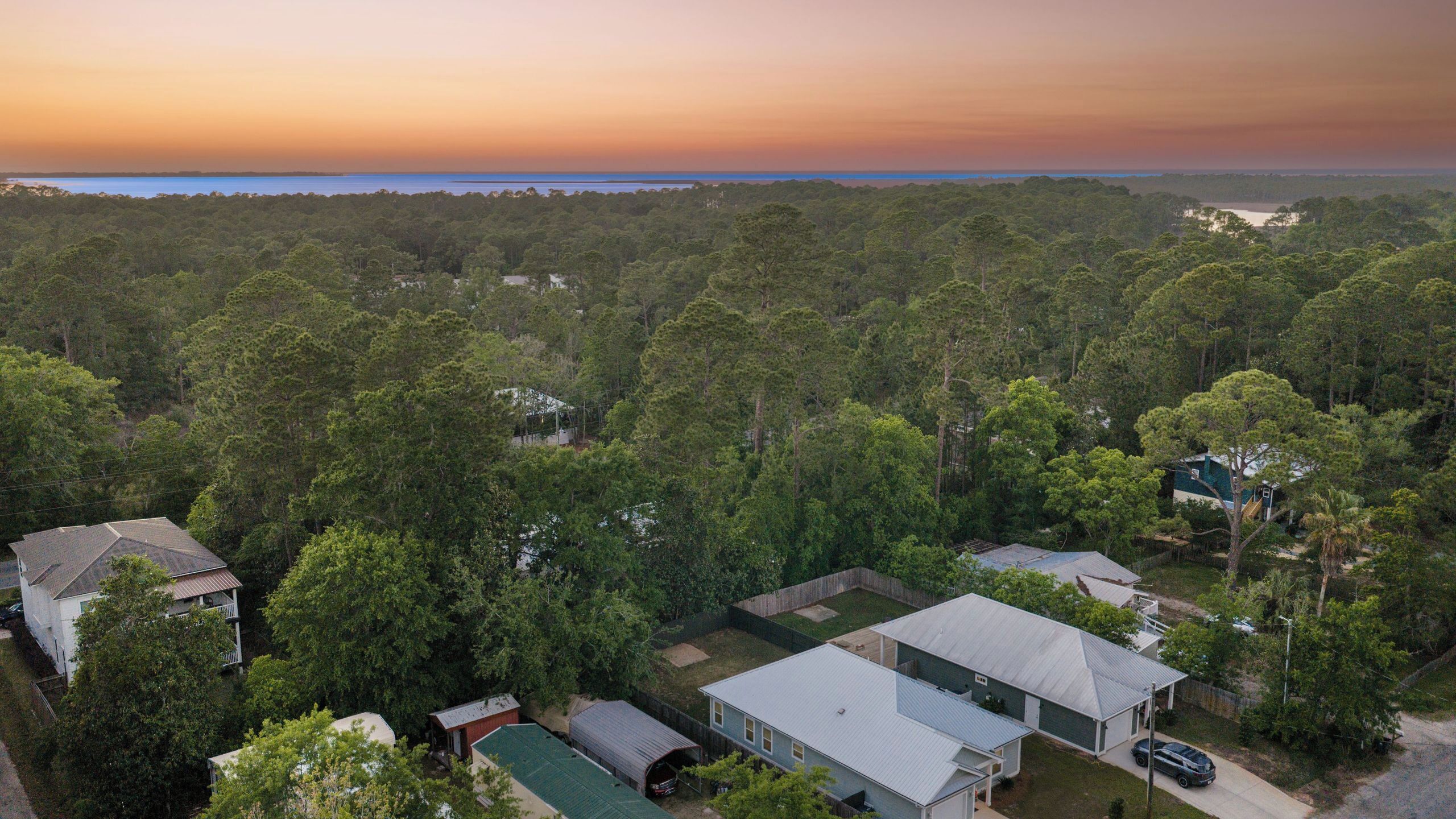 A residential area with houses surrounded by dense forest, a clear view of the sky, and a distant body of water.