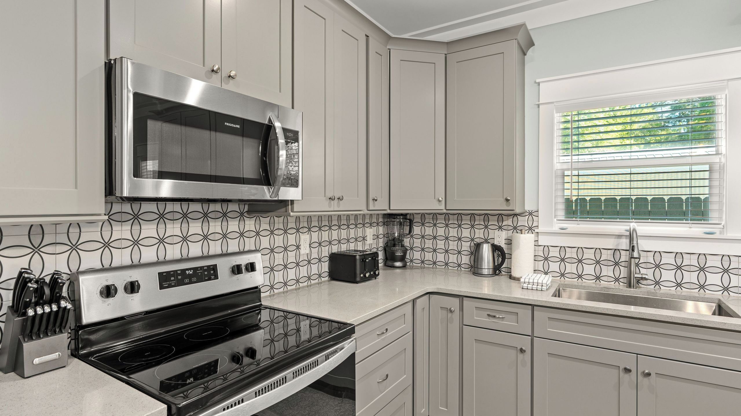 A modern kitchen with stainless steel appliances, a patterned backsplash, and a window with blinds.