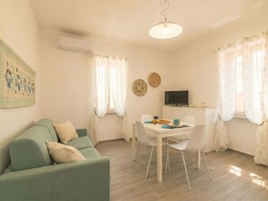 Ein Wohnzimmer mit einem grünen Sofa, einem Esstisch mit Stühlen, einem Fernseher auf einem Standfuß und einem Fenster mit Vorhängen.
