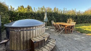 A hot tub with a wooden exterior, a wooden table with chairs, and a hedge in the background.