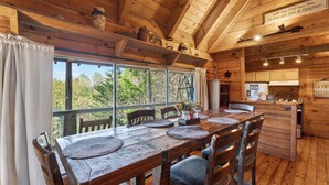 A wooden cabin interior with a dining table, chairs, a kitchen area, and large windows overlooking greenery. - Maggie's Cabin by Avantstay Hot Tub + Fire Pit + Mountain Views + All-wood Cabin Near National Park (Townsend)