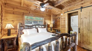 A log cabin bedroom with a bed, two bedside tables with lamps, a ceiling fan, and a window.