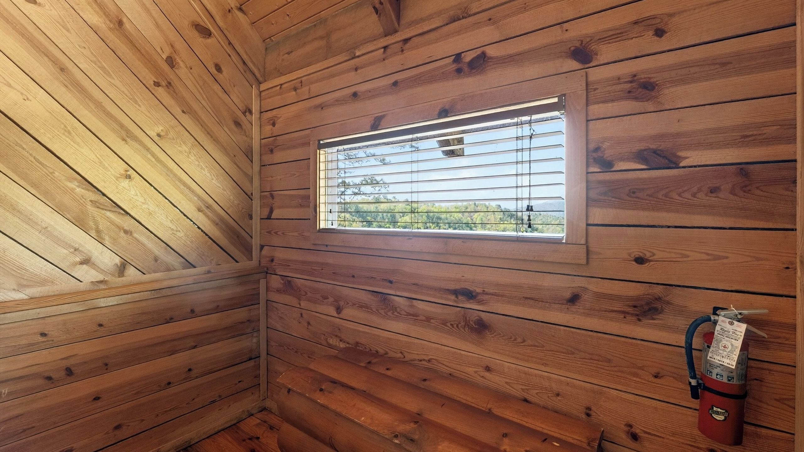 A wooden cabin room with a window, a fire extinguisher, and a wooden ceiling.