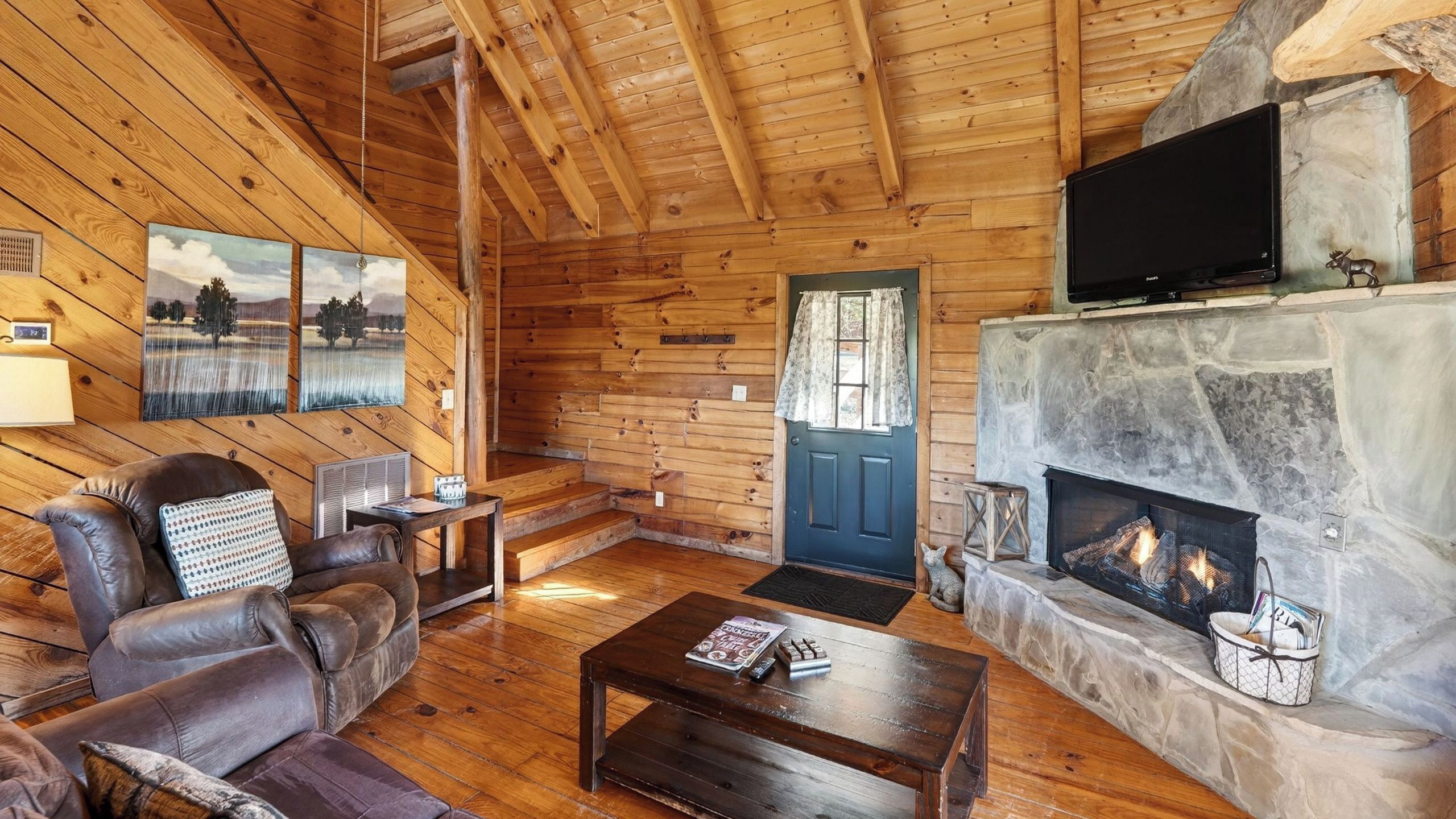 A cozy cabin interior with a fireplace, wooden walls, a leather sofa, and a television.