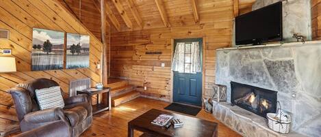 A cozy cabin interior with a fireplace, wooden walls, a leather sofa, and a television.