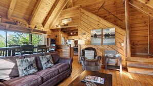 A wooden cabin interior with a sofa, armchair, dining table, and kitchen area. - Maggie's Cabin by Avantstay Hot Tub + Fire Pit + Mountain Views + All-wood Cabin Near National Park (Townsend)
