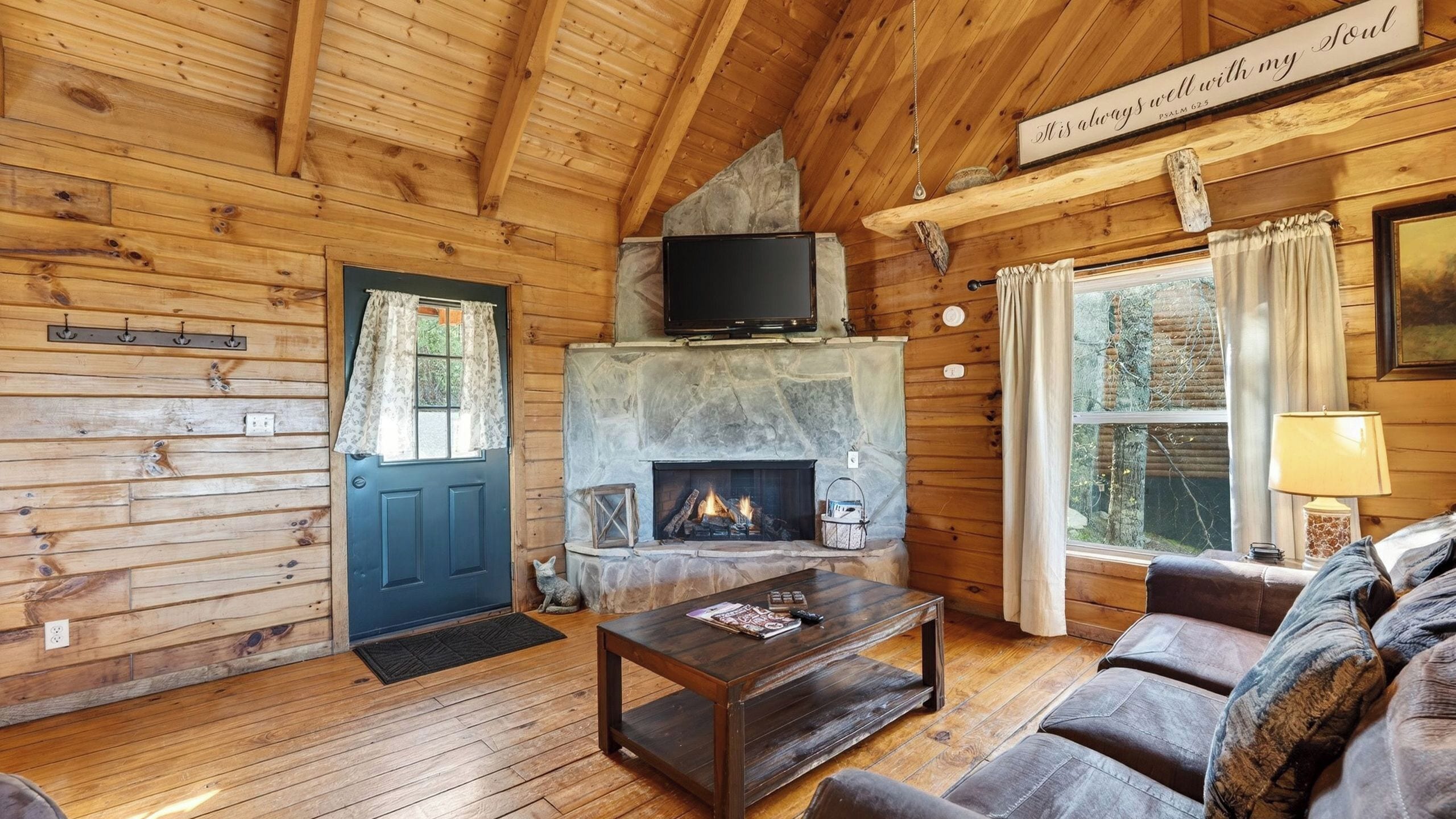 Ein gemütliches Blockhaus-Interieur mit Kamin aus Stein, Holzwänden, einem darüber montierten Fernseher, einer Couch und einem Fenster mit Vorhängen.