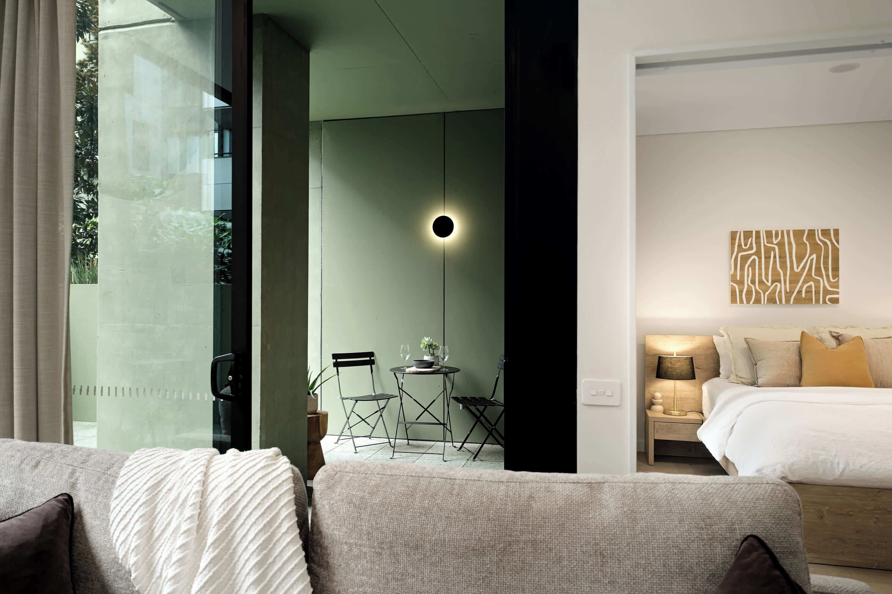 A modern hotel room with a bed, bedside table, lamp, and a view of the outdoors through a glass door.