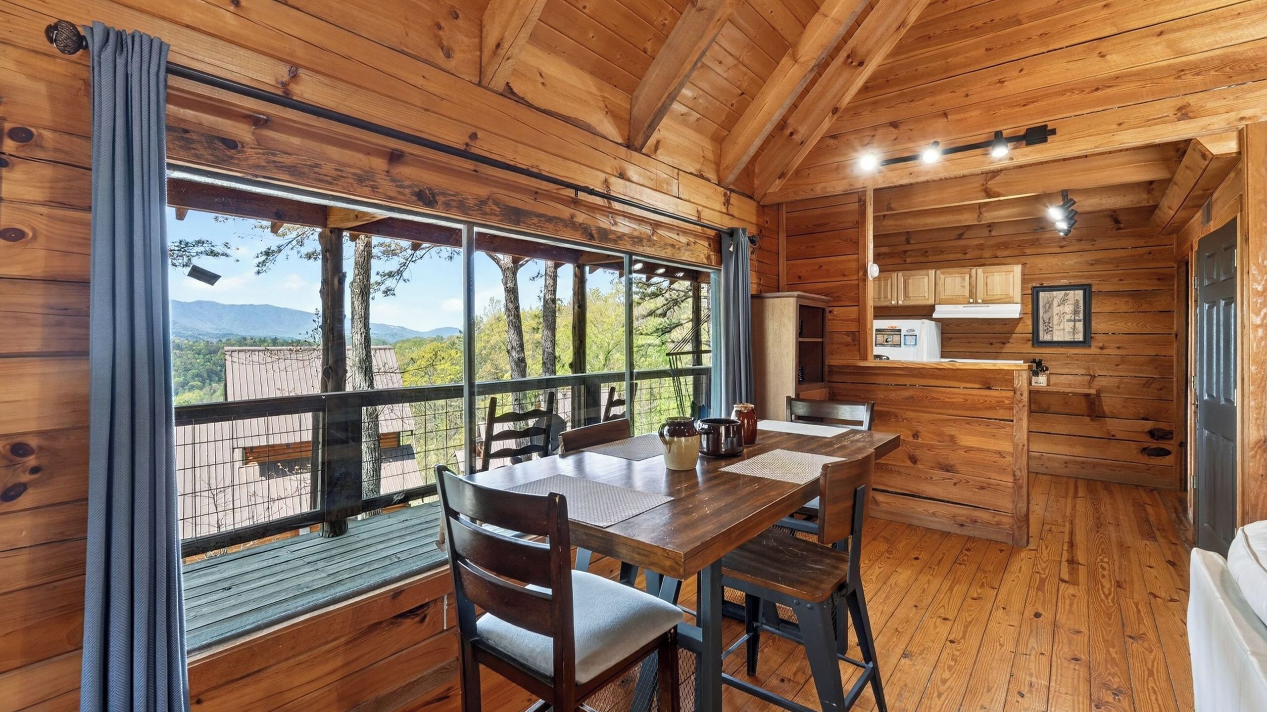 A wooden cabin interior with a dining table, chairs, a kitchen area, and large windows offering a view of the outdoors.