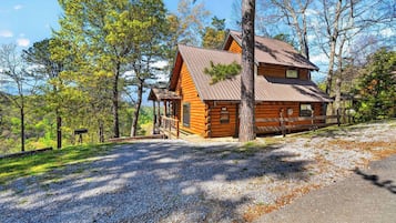 A log cabin with a porch, surrounded by trees and gravel.