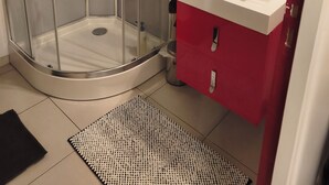 Une salle de bain moderne avec une douche à parois de verre, un lavabo blanc et des meubles rouges, ainsi qu’un petit tapis sur le sol.