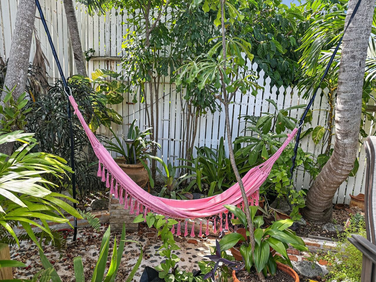 A hammock in a garden with various plants and a white fence.