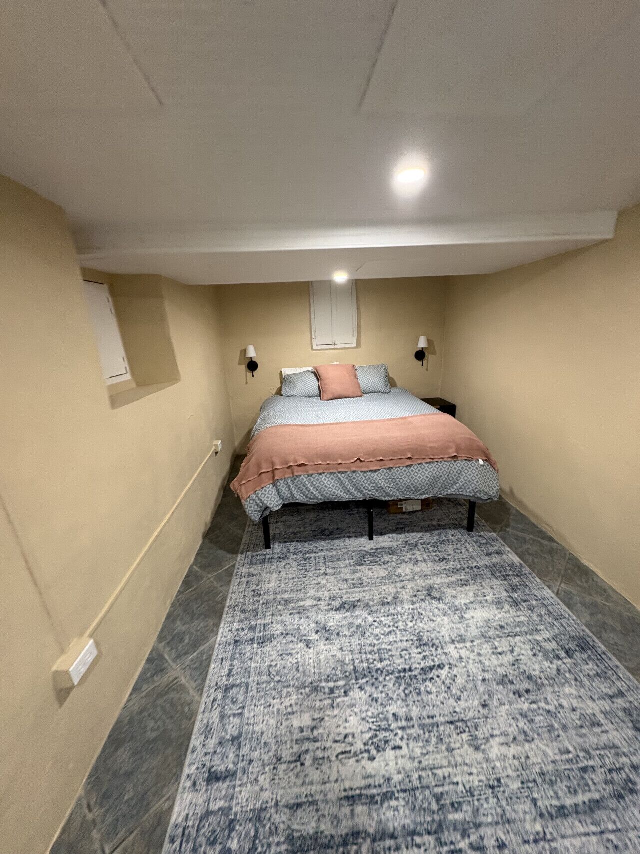 Een slaapkamer met een bed, een vloerkleed, een raam en een wandlamp.