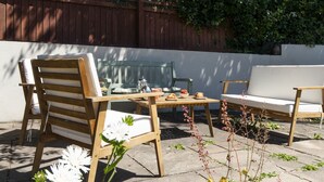Una terrazza esterna con mobili in legno, un tavolo con piano in vetro e un muro bianco sullo sfondo.