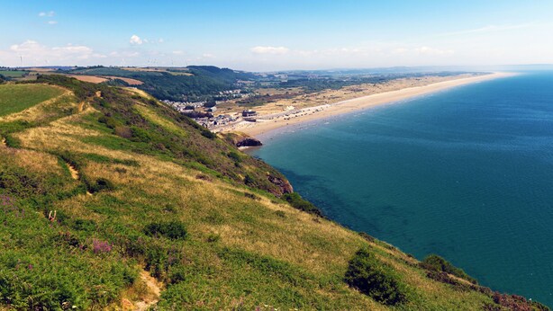 Paesaggio costiero con spiaggia, dolci colline e turbine eoliche in lontananza.