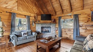 A log cabin interior with a stone fireplace, leather sofas, a wooden coffee table, and a flat-screen TV mounted above the fireplace.