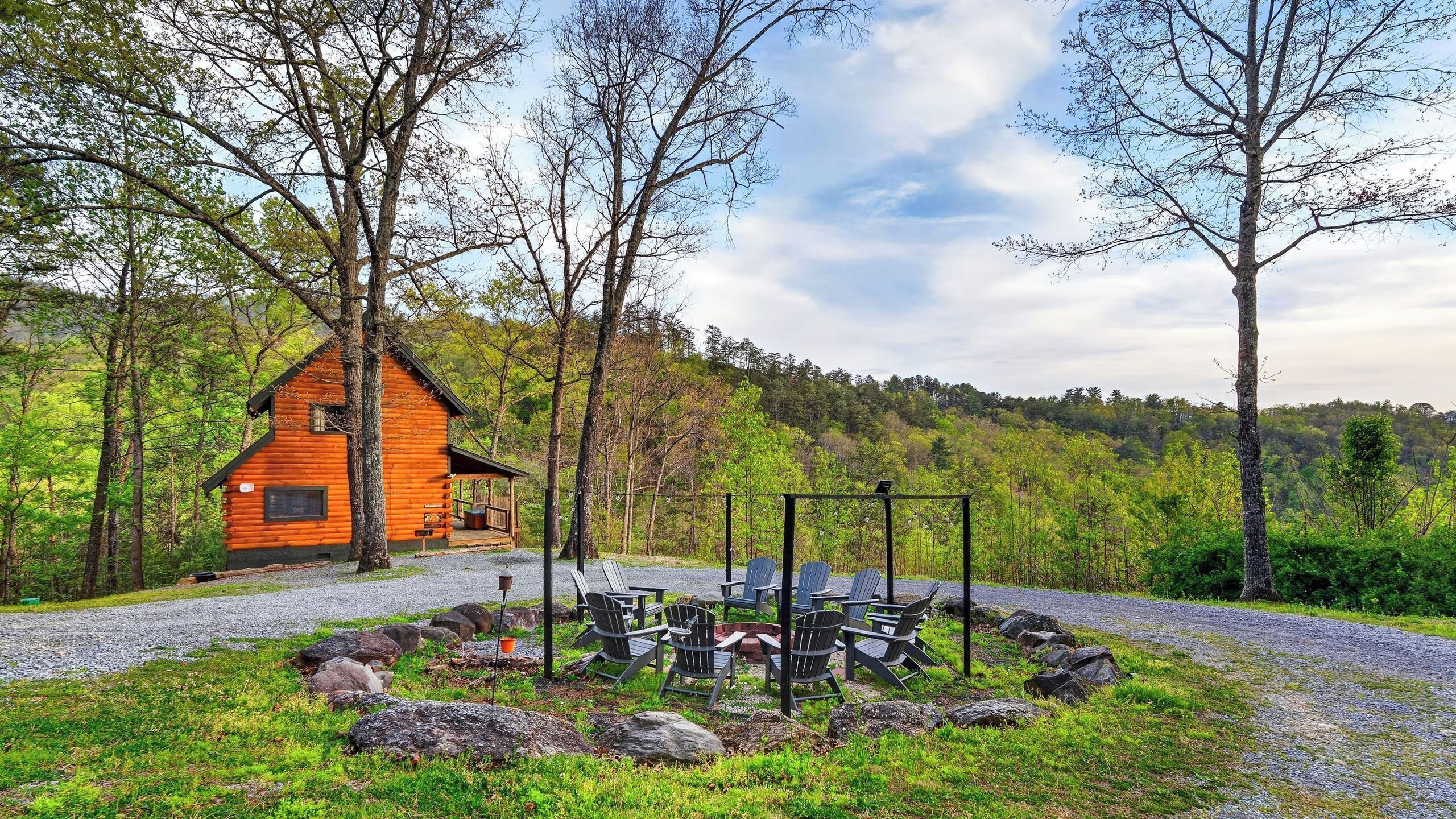 A log cabin with a porch, surrounded by a gravel path and a fire pit area with chairs.