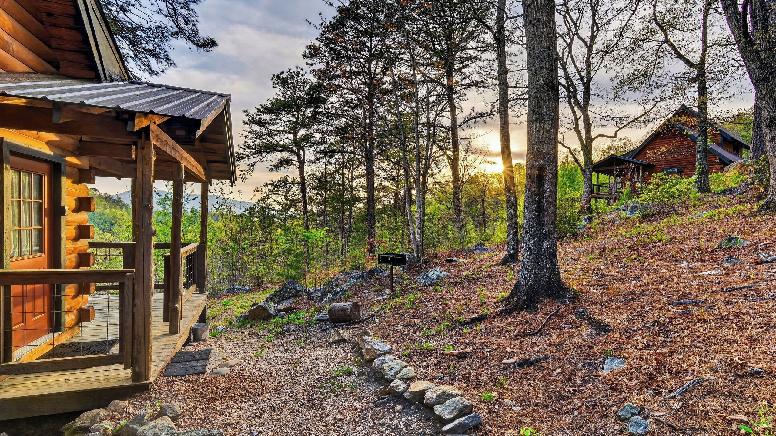 A rustic cabin with a wooden deck, surrounded by a forested area with a clear path leading to it.