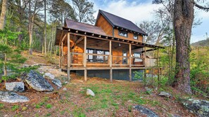 A cabin with a wooden exterior, a balcony, and a metal roof, surrounded by trees and rocks.