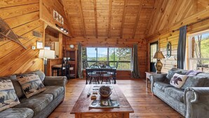 A wooden cabin interior with a dining table, two sofas, and a large window offering a view of the outdoors.