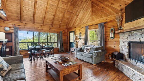 A wooden cabin interior with a dining area, sofa, fireplace, and large windows.