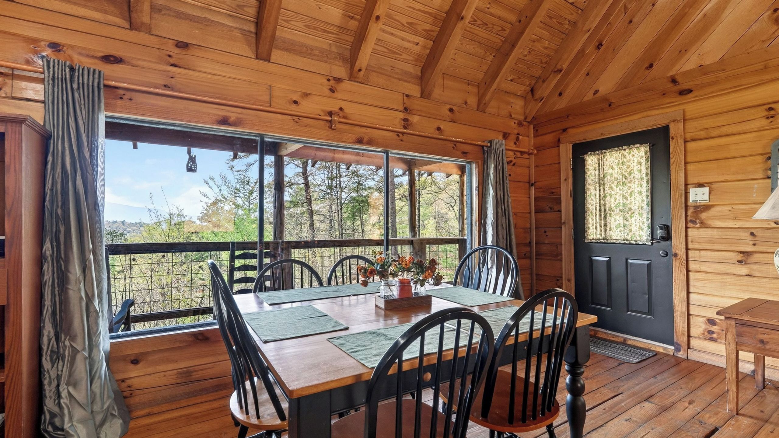 A wooden cabin with a dining area featuring a table and chairs, a black door, and large windows offering a view of the outdoors.