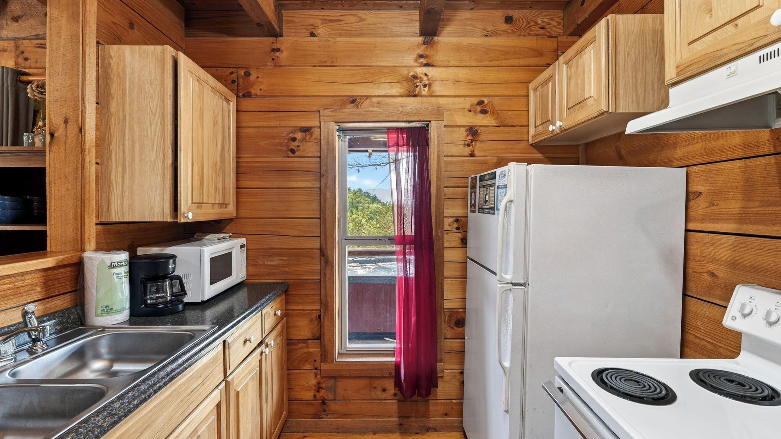 A compact kitchen with wooden walls, a window with a curtain, a white refrigerator, a stove, and a microwave.