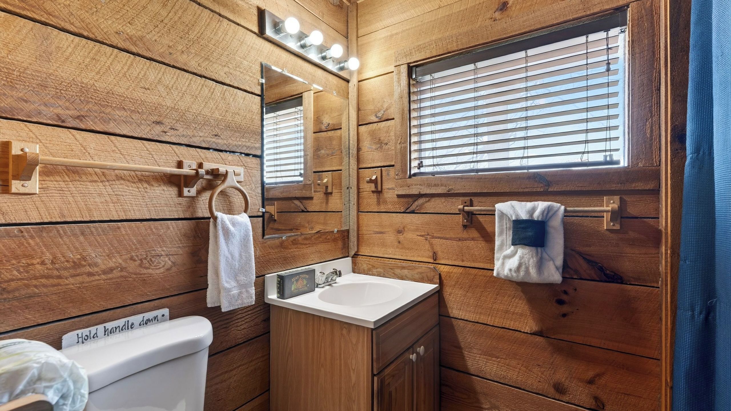 A wooden-paneled bathroom with a toilet, sink, mirror, towel rack, and window.