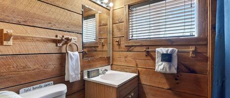 A wooden-paneled bathroom with a toilet, sink, mirror, towel rack, and window.
