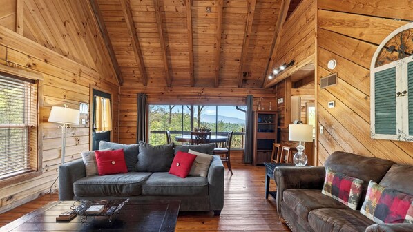A wooden cabin interior with a sofa, a coffee table, and a view through a large window.