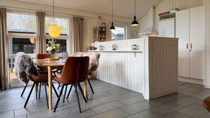 Una cocina moderna con zona de comedor, que presenta techo de madera, lámparas colgantes, refrigerador blanco y una mesa de comedor con sillas.