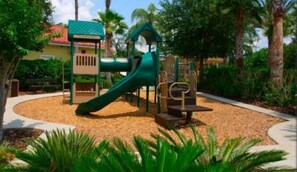 A playground with a slide and climbing structures, surrounded by a gravel area and palm trees. - Emerald Island Resort Double Beds Pet Friendly (Kissimmee)