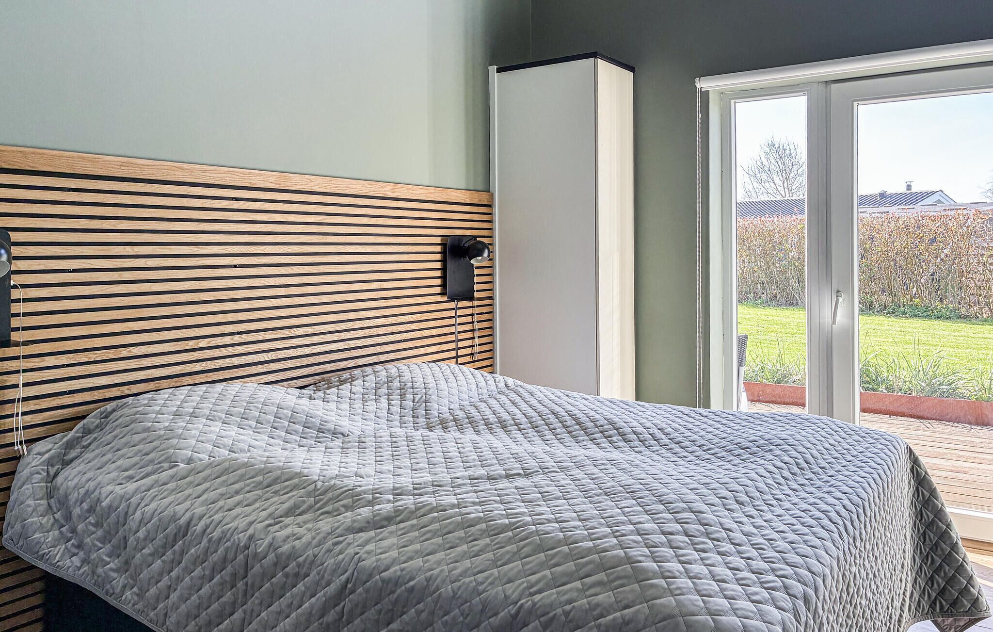 Une chambre avec un lit, une tête de lit en lattes de bois, une lampe fixée au mur et une porte-fenêtre coulissante donnant sur un balcon.