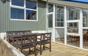 A wooden deck with a table and chairs, a building with large windows, and a view of the interior. - Pet friendly home in Hvide Sande with sauna (Hvide Sande)