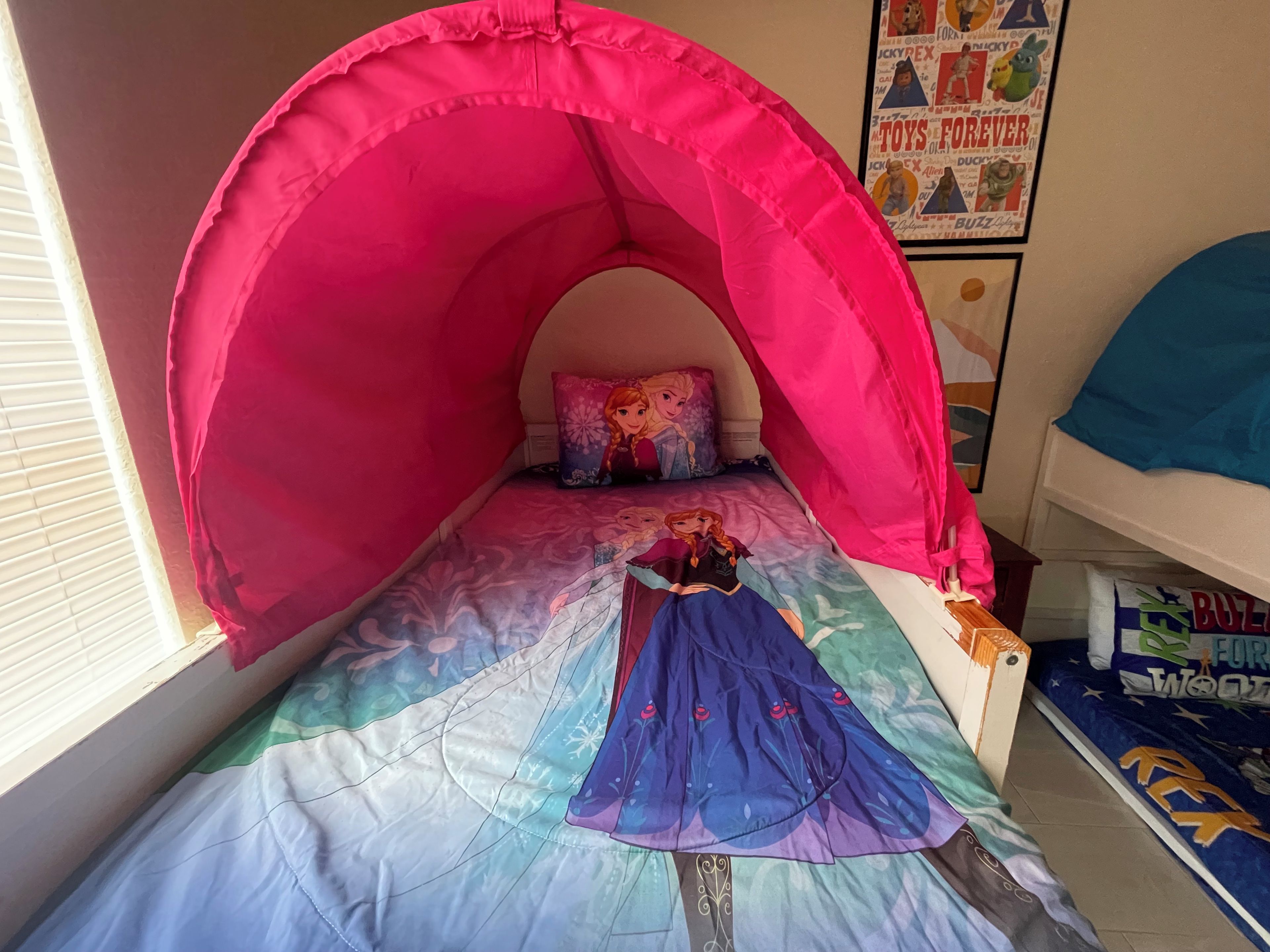 Una camera da letto con un letto a baldacchino rosa, una coperta a tema principessa Disney e un poster di un giocattolo appeso alla parete.