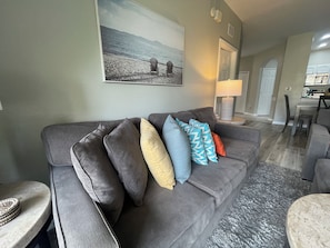 Ein Wohnzimmer mit grauem Sofa, mehreren Kissen, einem gerahmten Bild an der Wand und einer Lampe auf einem Beistelltisch.