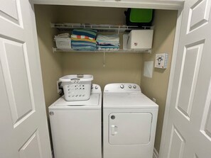 Une buanderie équipée de deux machines à laver et deux sèche-linge blancs, d’un panier et d’une étagère où sont pliés des vêtements et rangés les produits de lessive.