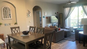 Ein Essbereich mit Holztisch und Stühlen, ein Wohnzimmer mit Sofa und Fernseher sowie ein Balkon mit Vorhängen.
