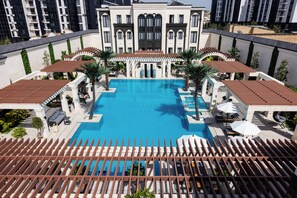 Uma piscina grande com pergolado, palmeiras e um edifício com janelas em arco ao fundo.