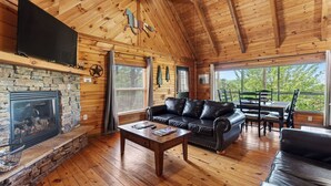 A wooden cabin interior with a stone fireplace, leather sofa, wooden dining table, and large windows. - Sónyah's Cabin by Avantstay Hot Tub + Deck + Firepit + Mountain Views Mins to Downtown (Townsend)