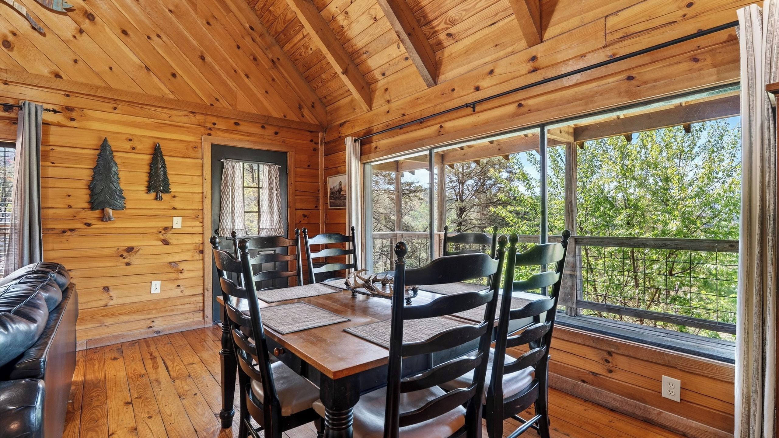 A wooden cabin interior with a dining table, chairs, a leather couch, and large windows offering a view of the outdoors.